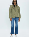 A person stands against a plain background wearing the Pistola Elva Onion Stitch Puffer Jacket in military green over a black top, paired with blue flared jeans featuring slight distressing at the knees, white sneakers, and dark sunglasses. They have long dark hair and stand with a relaxed posture.