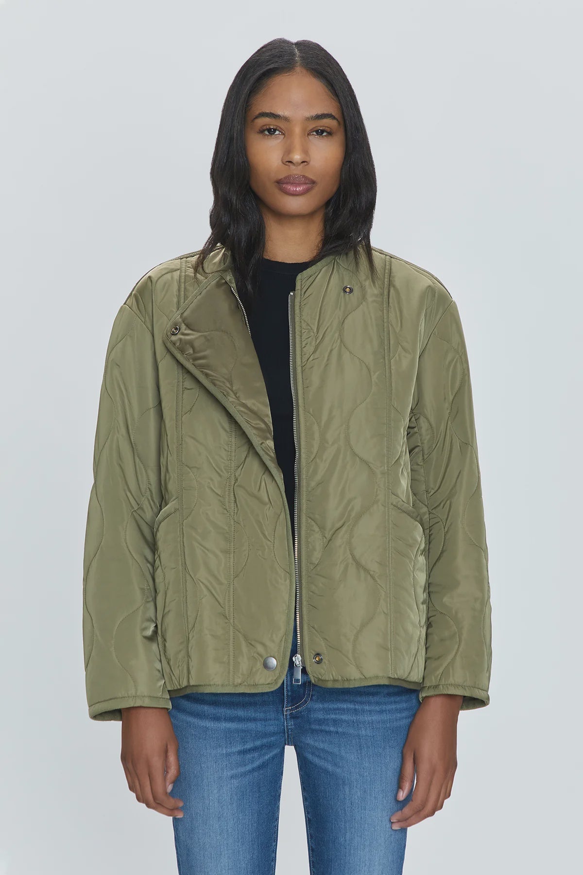 A woman with long, dark hair is standing against a plain, light background. She is wearing the Elva Onion Stitch Puffer Jacket by Pistola in military green over a black top and blue jeans, looking directly at the camera with a neutral expression.