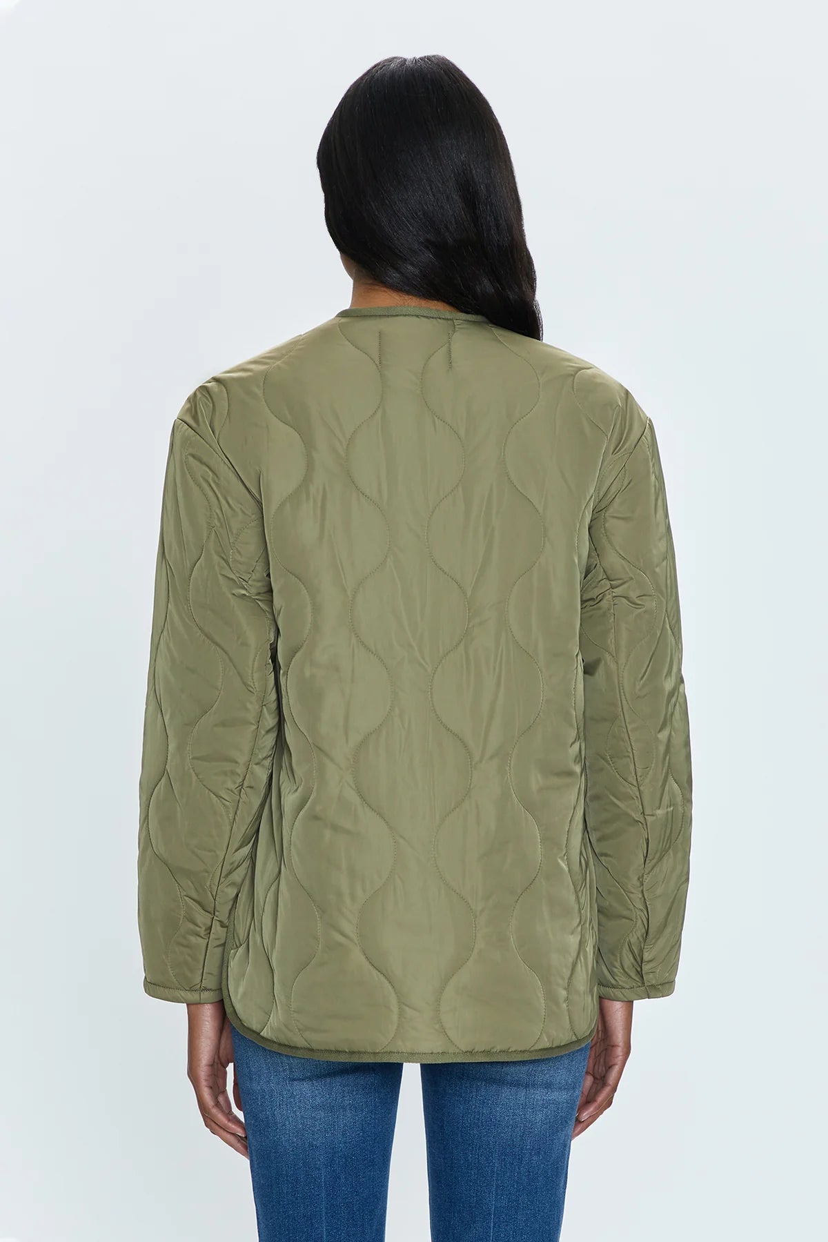 A person with long dark hair is seen from behind, wearing the Pistola Elva Onion Stitch Puffer Jacket in military green and blue jeans. The jacket showcases a simple, onion stitch quilting pattern against a plain, light gray backdrop.