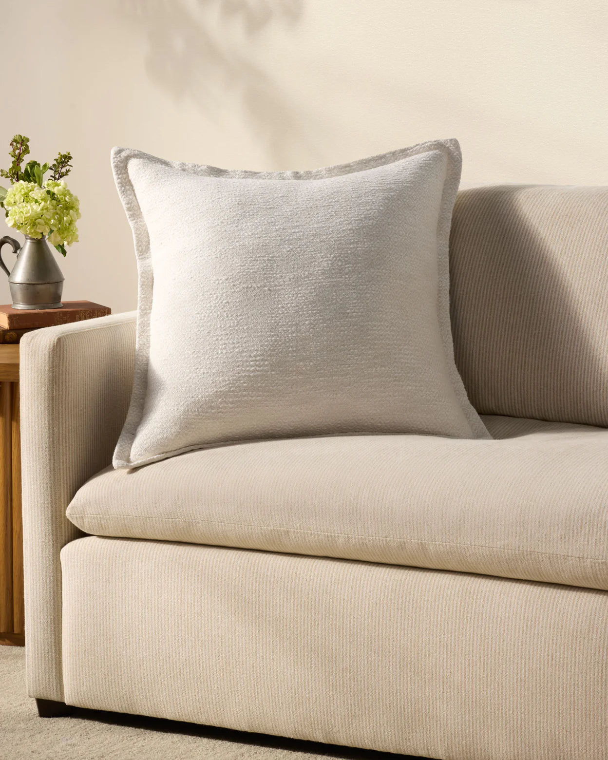 A light beige sofa is accented with the Loloi Rugs PCJ0022 Pillow 22x22 in textured light gray, set against a cream wall. Beside it, a small wooden side table with an antique bronze finish displays a vase of green and white flowers.