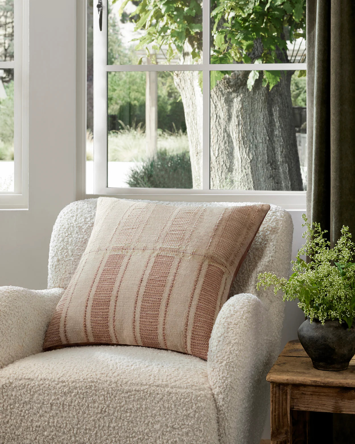 A cozy white boucle armchair sits by a sunlit window, featuring the Marin Natural 22x22 pillow by Loloi Rugs in beige and pink plaid. Outside, trees and greenery shine, while a small wooden table with a leafy plant rests nearby.