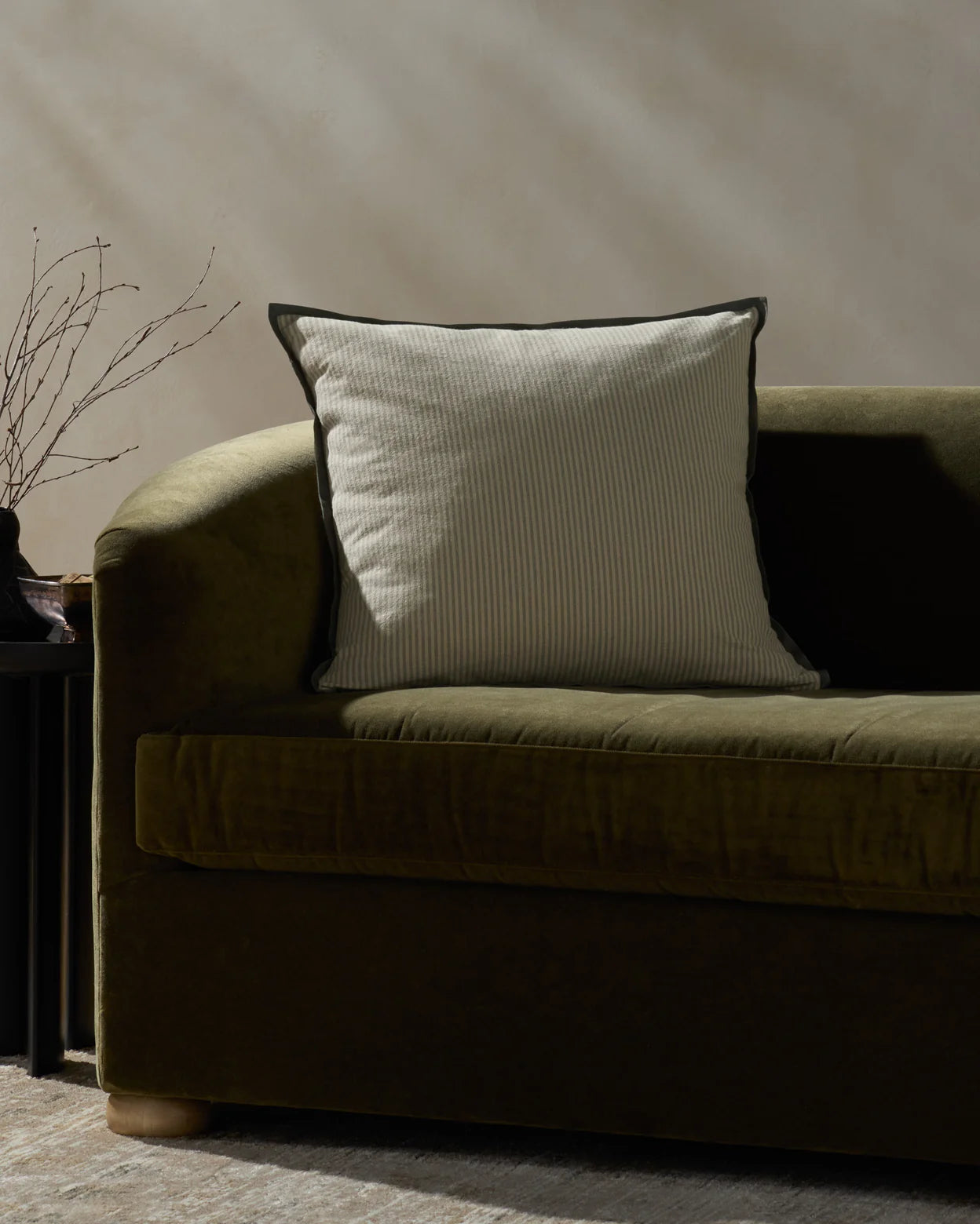 A green velvet sofa with the Loloi Rugs PJBR0001 Natural BB Pillow, a light beige striped cushion with a hidden zipper, sits by a black side table holding bare branches, set against a neutral wall and rug.