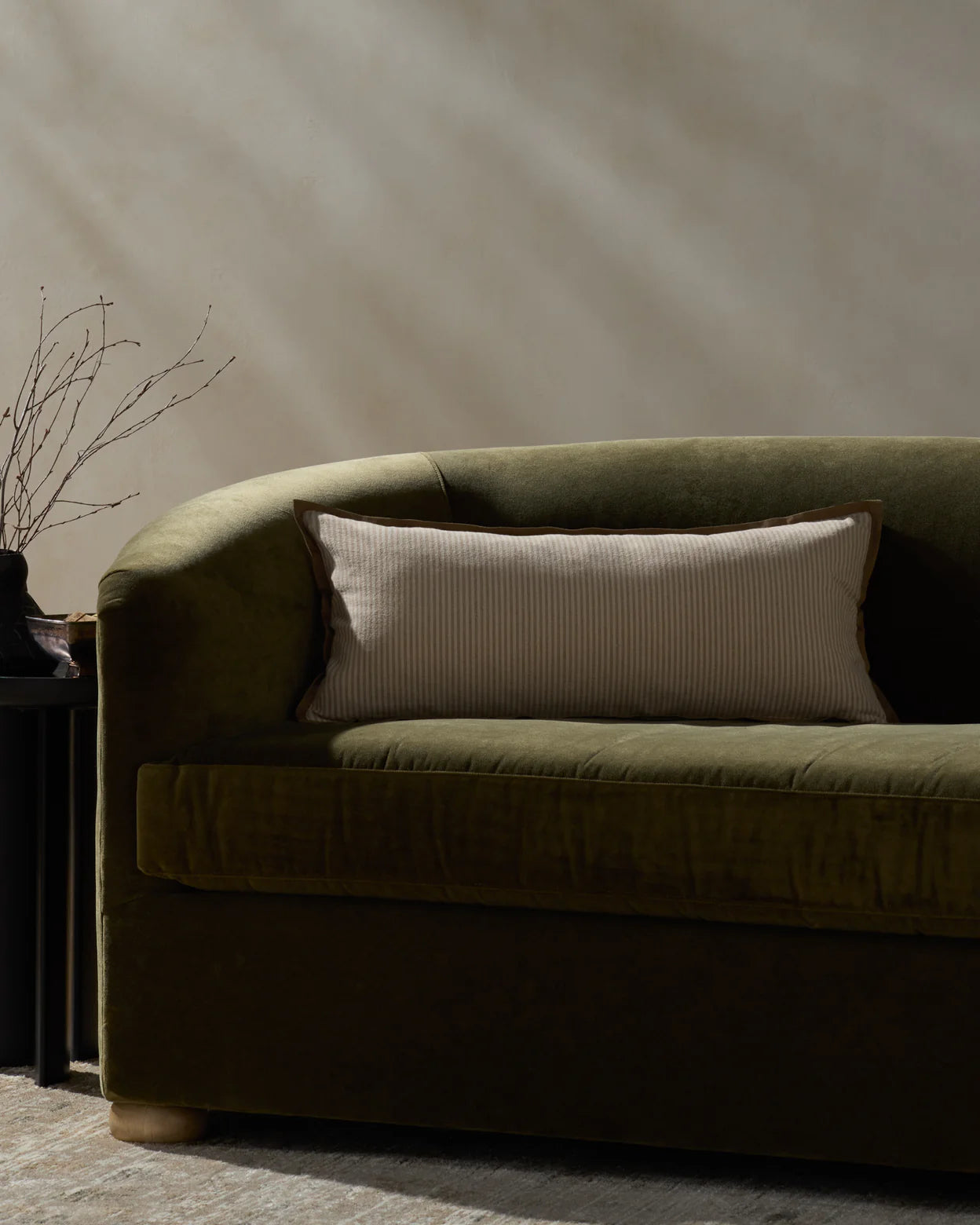 An olive green velvet sofa with a Loloi Rugs PJBR0001 Natural BR Pillow, a rectangular beige cotton cushion with a hidden zipper, sits next to a small black side table holding a vase of bare branches. Soft natural light casts shadows on the neutral wall.