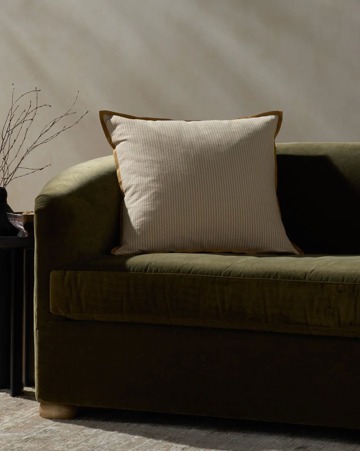 The PJBR0001 Natural GO Pillow by Loloi Rugs, featuring cream and beige stripes with brown piping and a hidden zipper, rests on an olive green velvet sofa beside a black side table topped with a minimalist vase of bare branches.