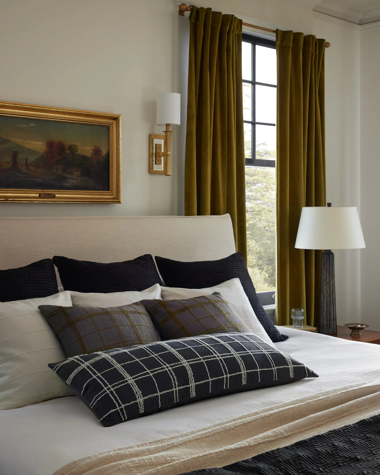 A cozy bedroom with a beige upholstered headboard, Loloi Rugs PJS0012 Graphite/Ivory Pillow (13x35), layered black and plaid pillows, a landscape painting above the bed, olive green curtains, a window, and a lamp on a wooden nightstand.