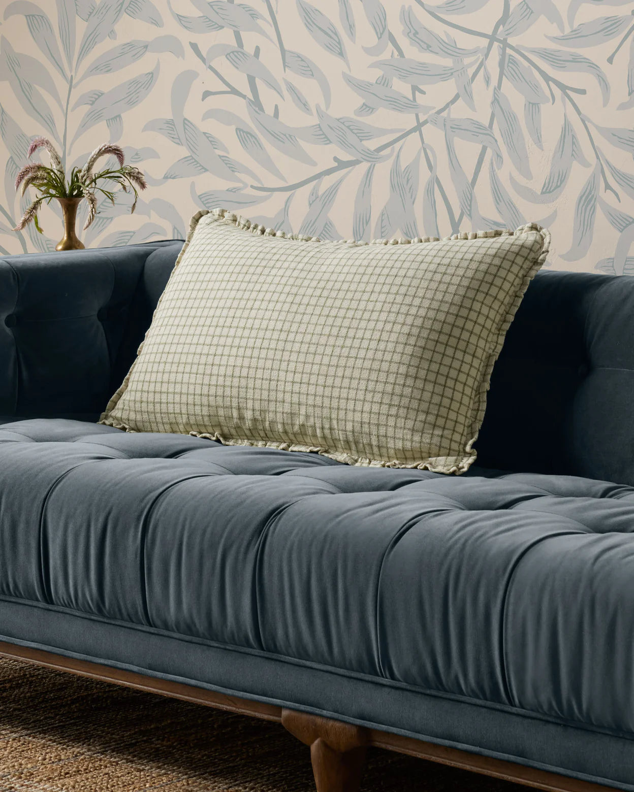 The Loloi Rugs PCJ0012 Ivory Moss pillow, featuring a light green checkered design with ruffle edges and a hidden zipper, rests on a dark blue velvet tufted sofa against beige and blue leafy wallpaper. A vase with flowers sits nearby.