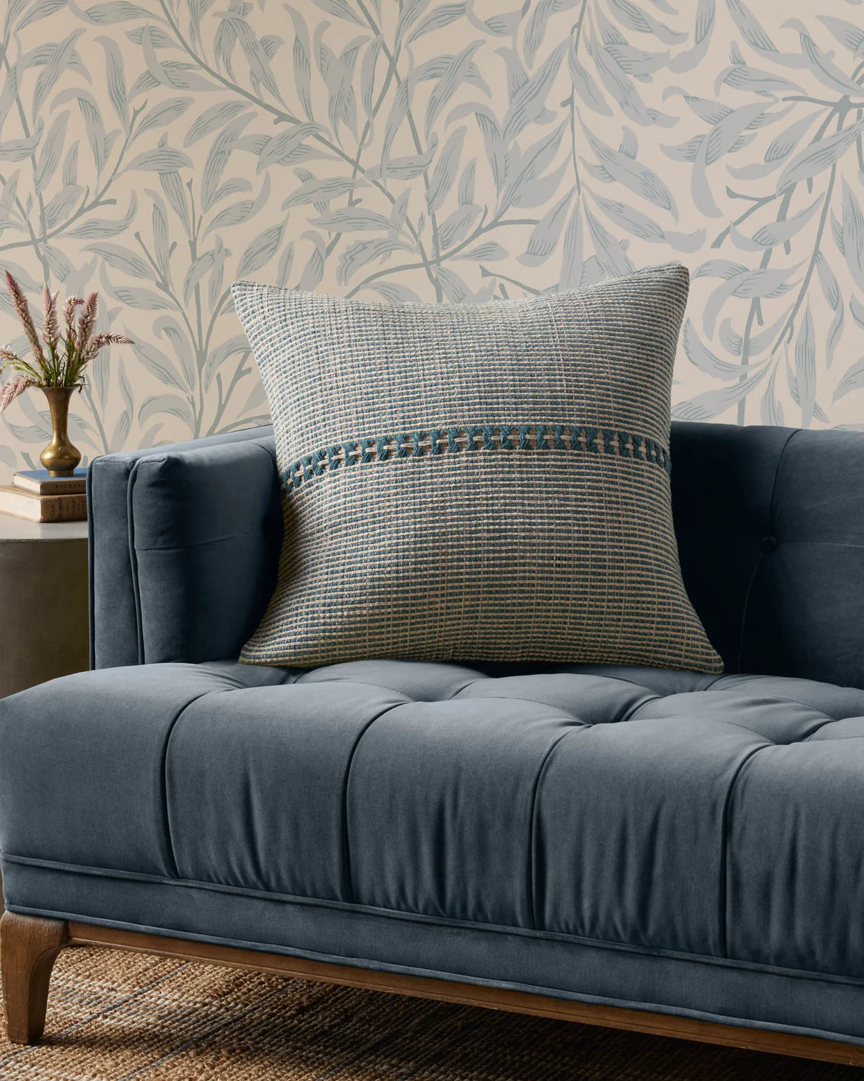 The PCJ0018 Blue Natural sofa by Loloi Rugs features tufted blue polyester, wooden legs, and a large woven cotton pillow with a blue-green stripe. It sits beside leafy wallpaper and an antique bronze side table with a vase of flowers.