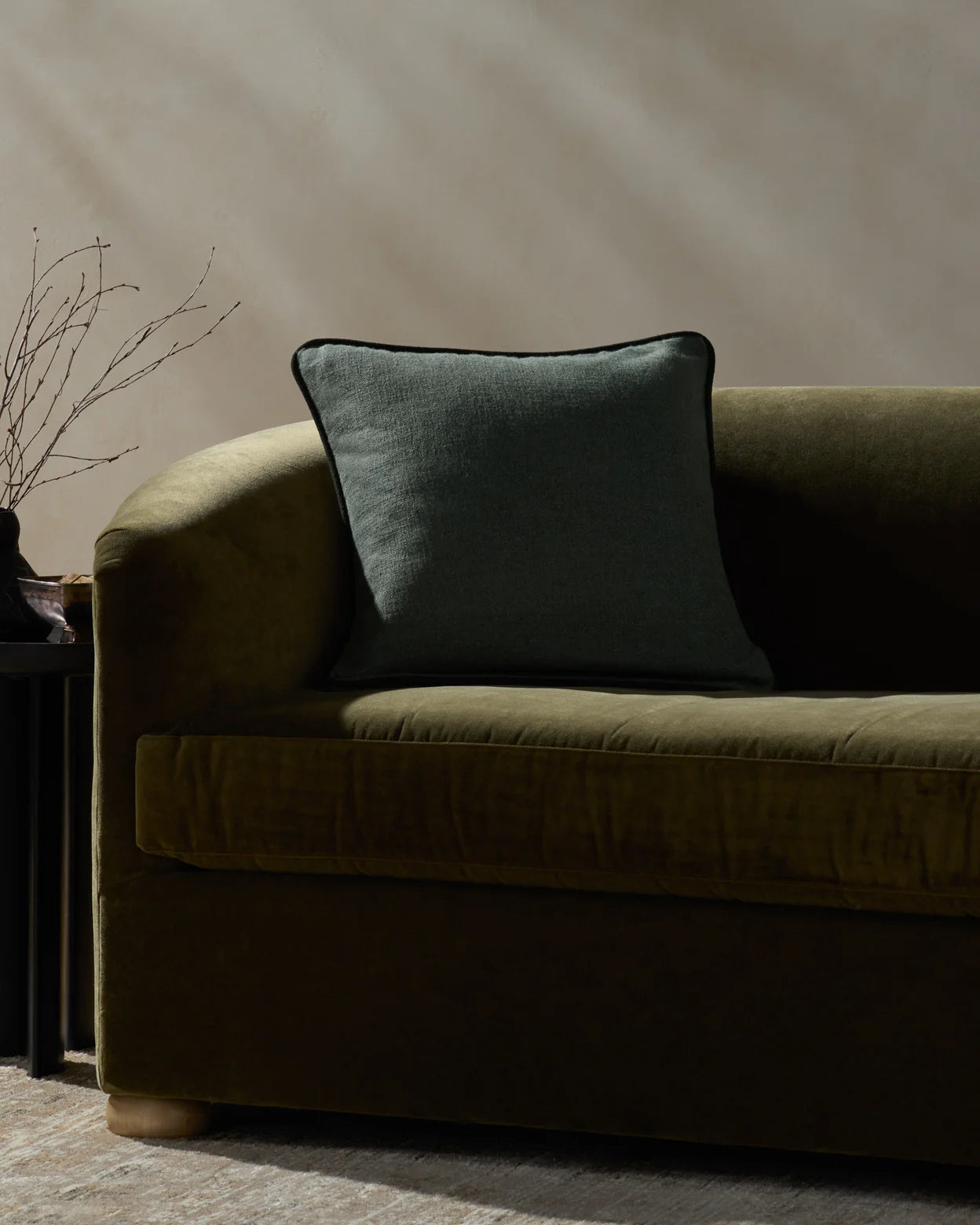 A dark green velvet sofa with a Loloi Rugs Hudson Blue Pillow 20x20 sits next to a small black side table holding a vase with thin branches. Sunlight casts soft shadows on the beige wall behind.