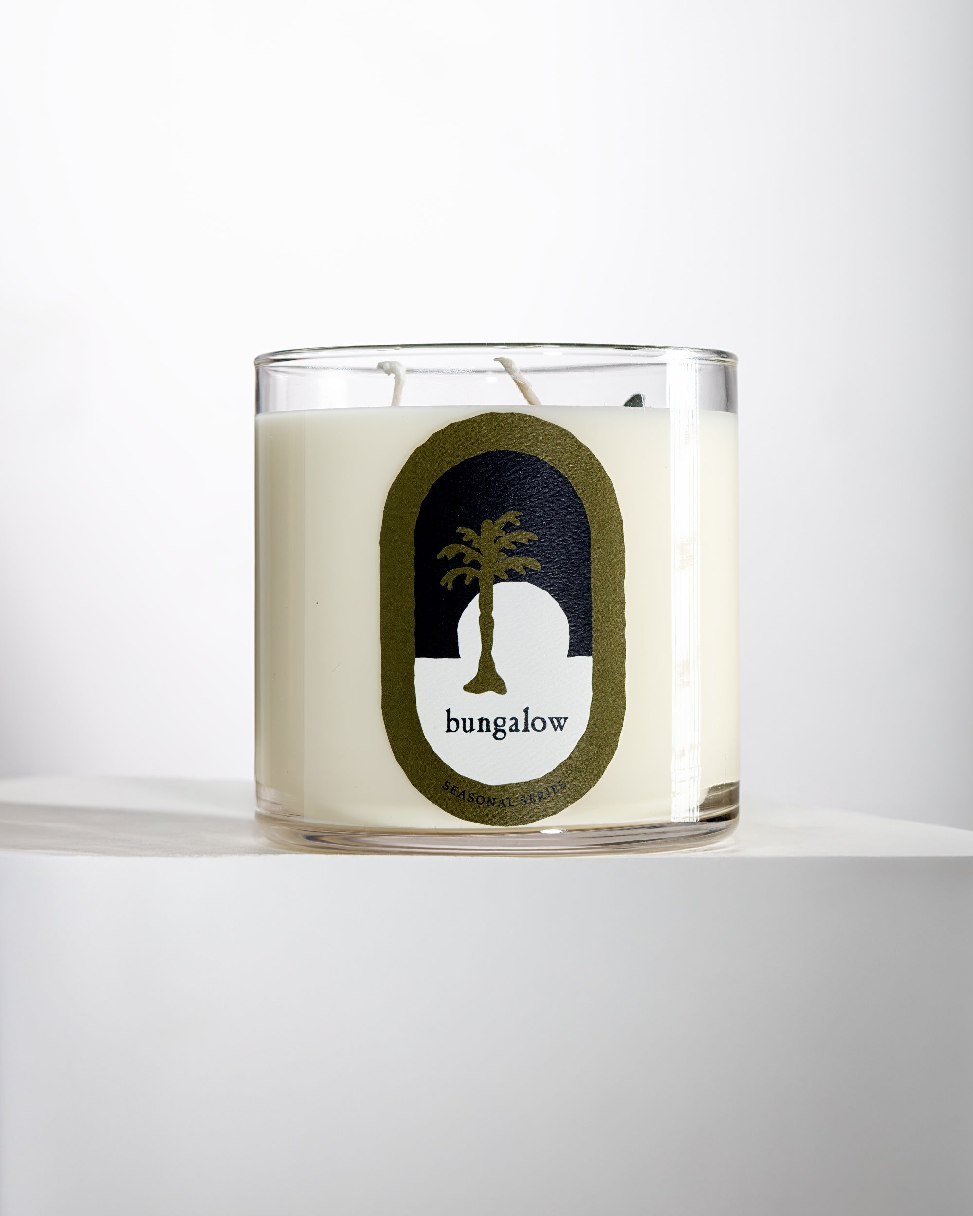 The Mame Signature Candle WINTER is a white holiday candle in a clear glass jar featuring a palm tree silhouette with the word "bungalow"—a perfect choice for cozy winter evenings.