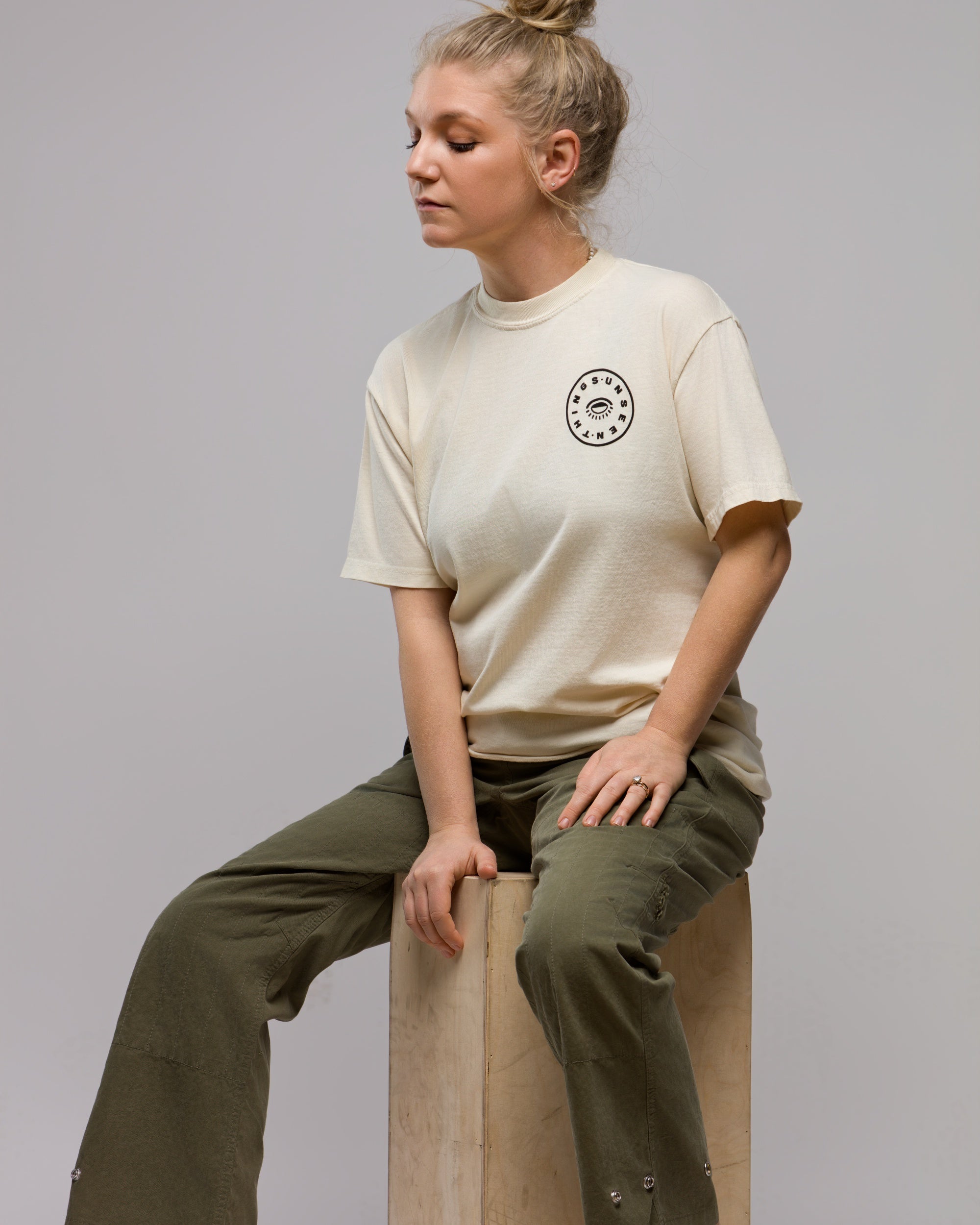 A woman with blonde hair in a bun sits on a wooden block, wearing the Things Unseen T-Shirt by Things Unseen with a small chest logo and olive green pants, gazing calmly to the side.