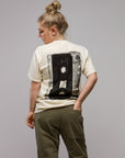 A person with blond hair in a messy bun faces away, wearing the Things Unseen T-Shirt in cream ivory with a bold black vintage cassette graphic on the back and olive green pants, standing against a plain gray background.