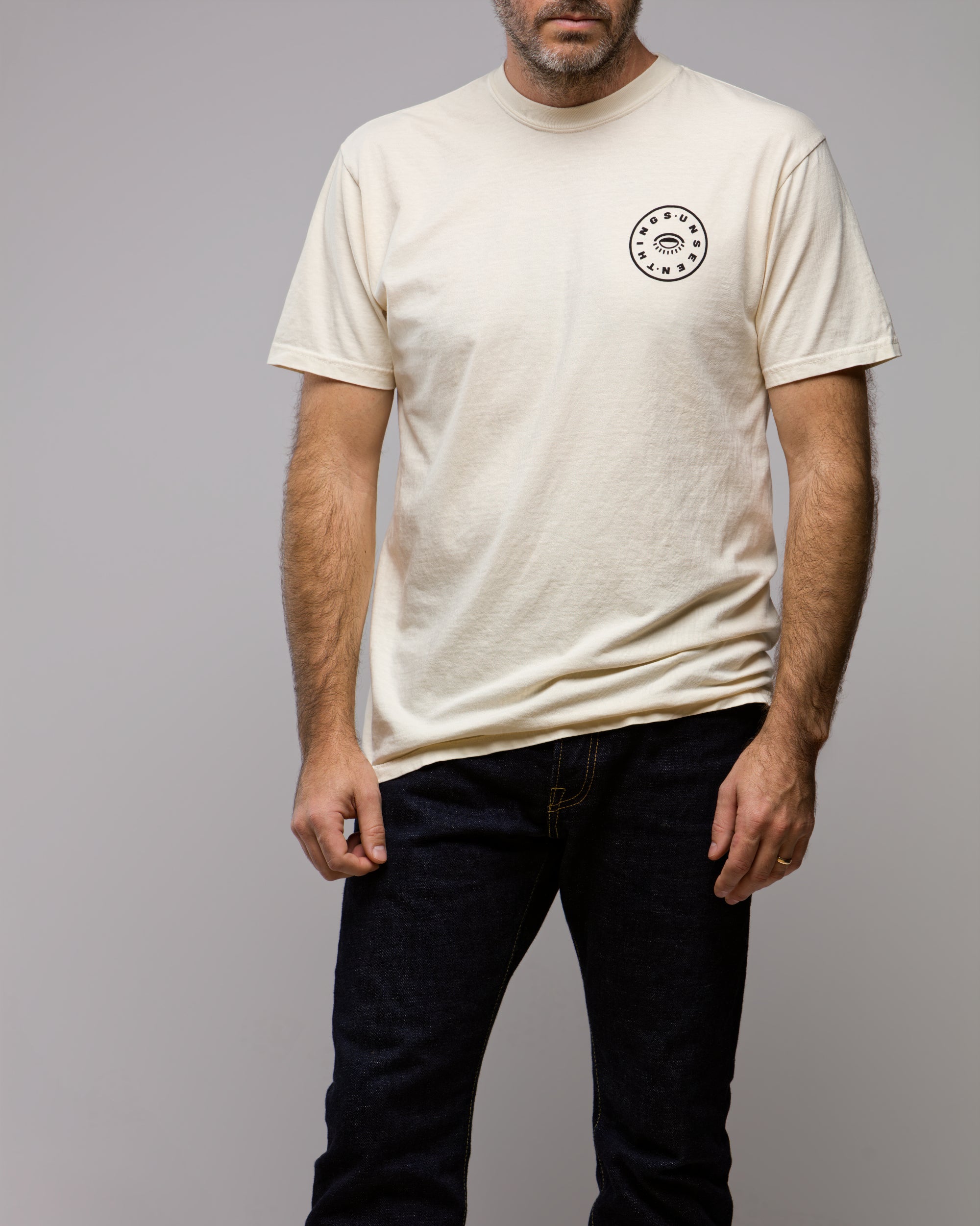 A man models the Things Unseen T-Shirt by Things Unseen, featuring a cream ivory color and a small round logo on the left chest, paired with dark jeans against a plain light gray background. His face is partially out of frame.