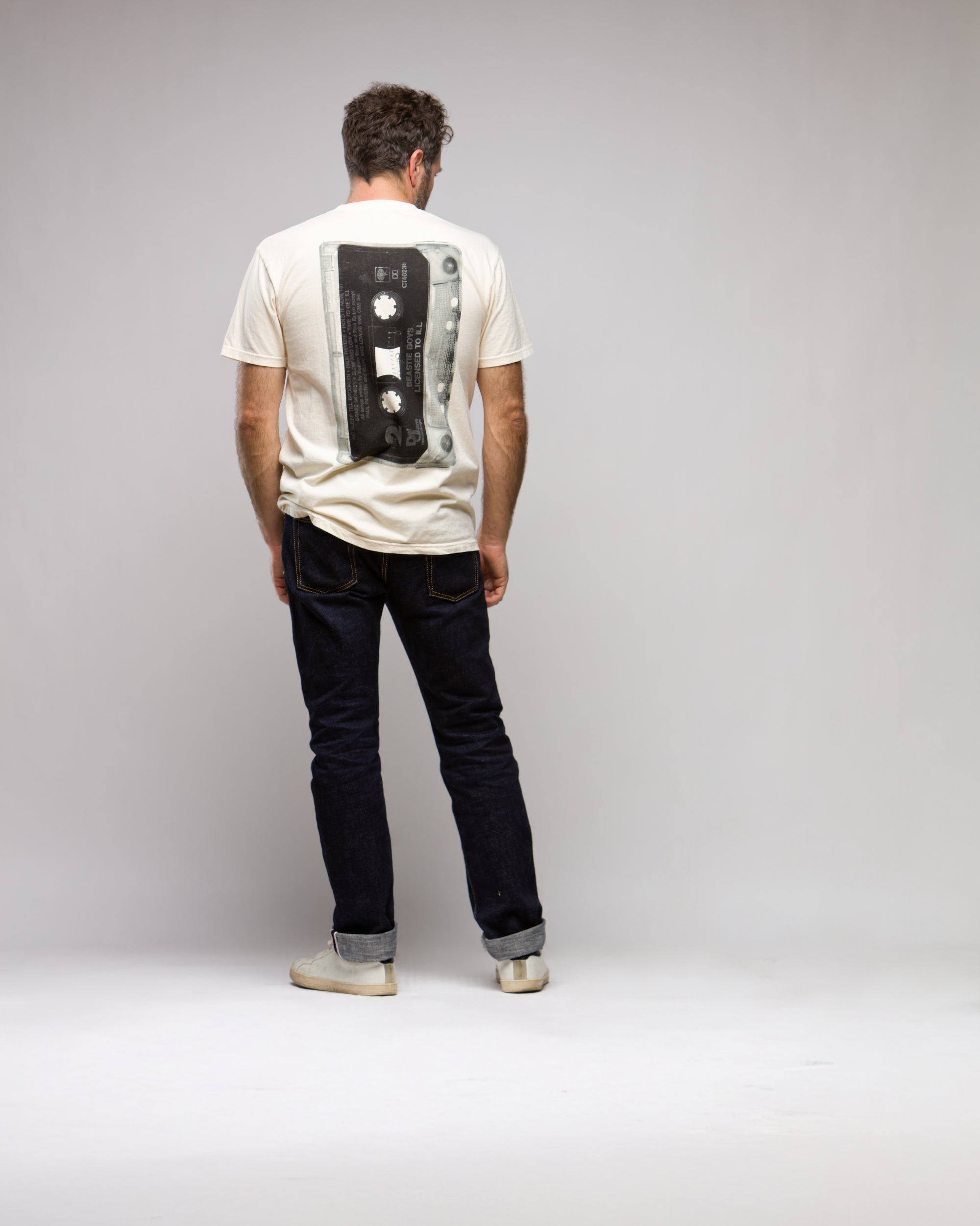 A man stands facing away in the Things Unseen T-Shirt by Things Unseen, featuring a vintage cassette design inspired by the Beastie Boys' License to Ill album. He pairs it with dark jeans and white shoes, set against a plain light gray background.