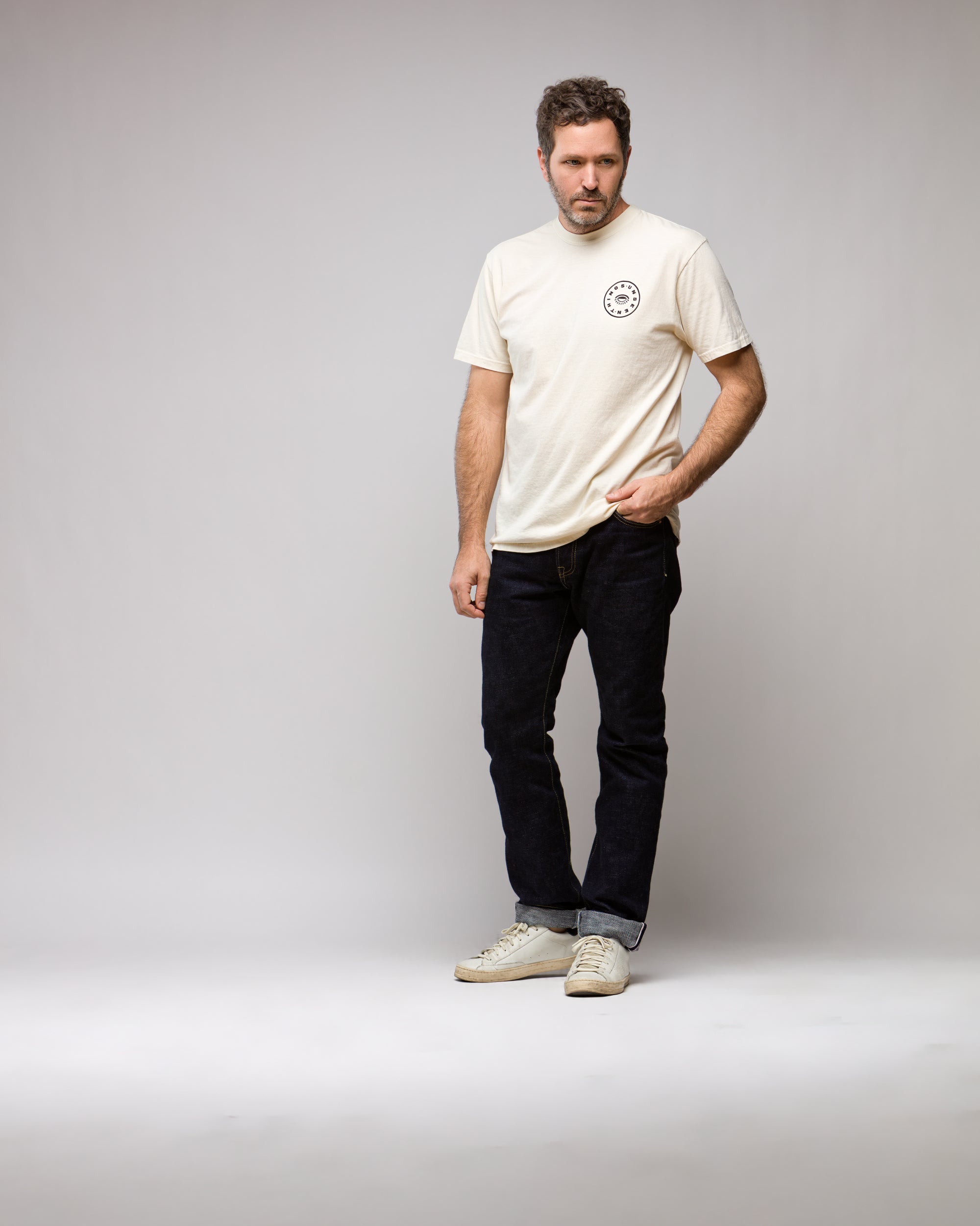 A man with short, curly hair and a beard stands in a studio wearing the Things Unseen T-Shirt by Things Unseen—cream ivory with a small chest logo—paired with dark jeans and white sneakers, looking slightly left with a neutral expression.