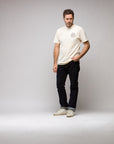 A man with short, curly hair and a beard stands in a studio wearing the Things Unseen T-Shirt by Things Unseen—cream ivory with a small chest logo—paired with dark jeans and white sneakers, looking slightly left with a neutral expression.