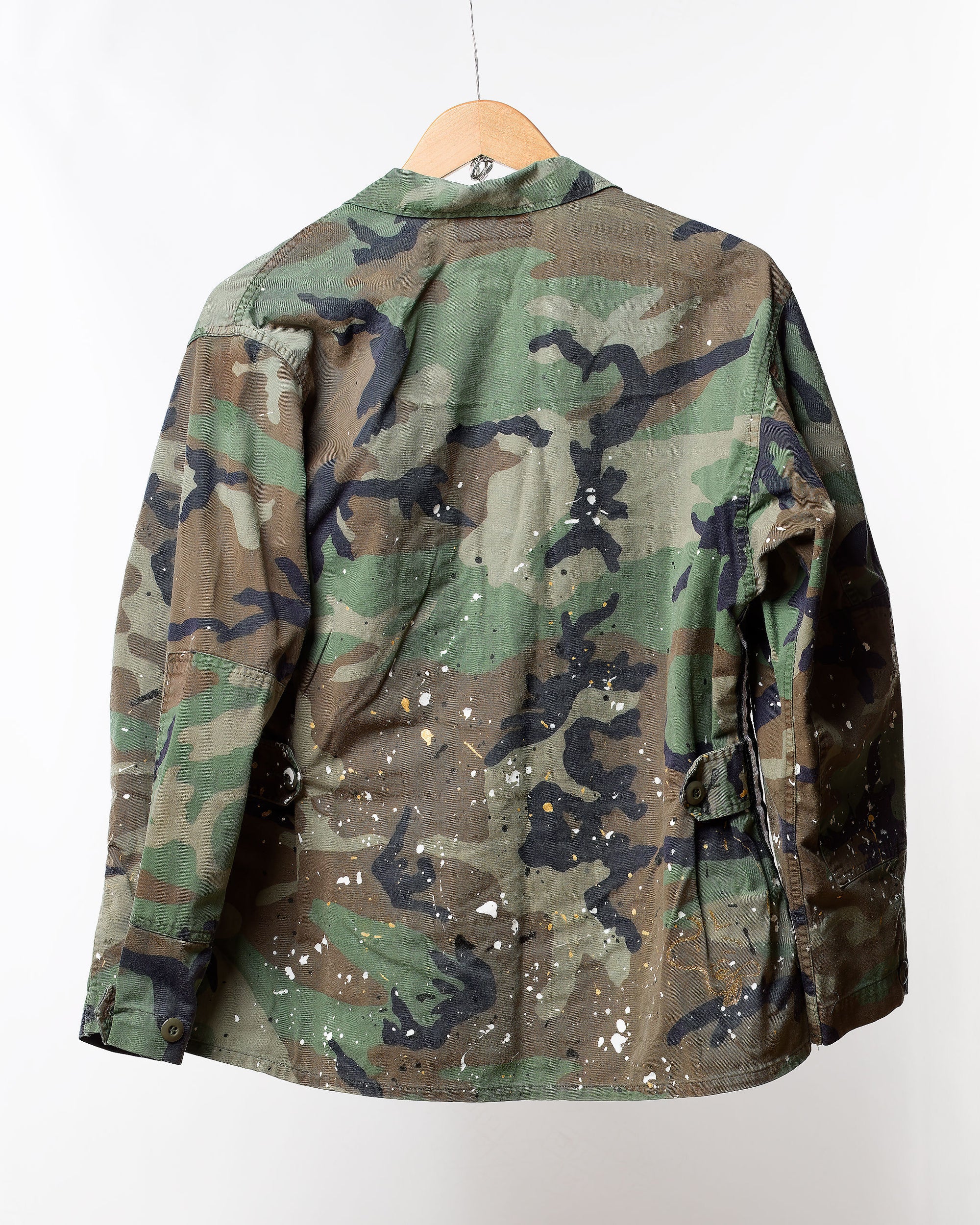 The Camo Jacket with Splatter & Trim by Tak Design is a vintage military camo piece that showcases a creative individual touch. It features splattered paint and hangs on a wooden hanger against a white background, highlighted by unique shades of green, brown, and black.