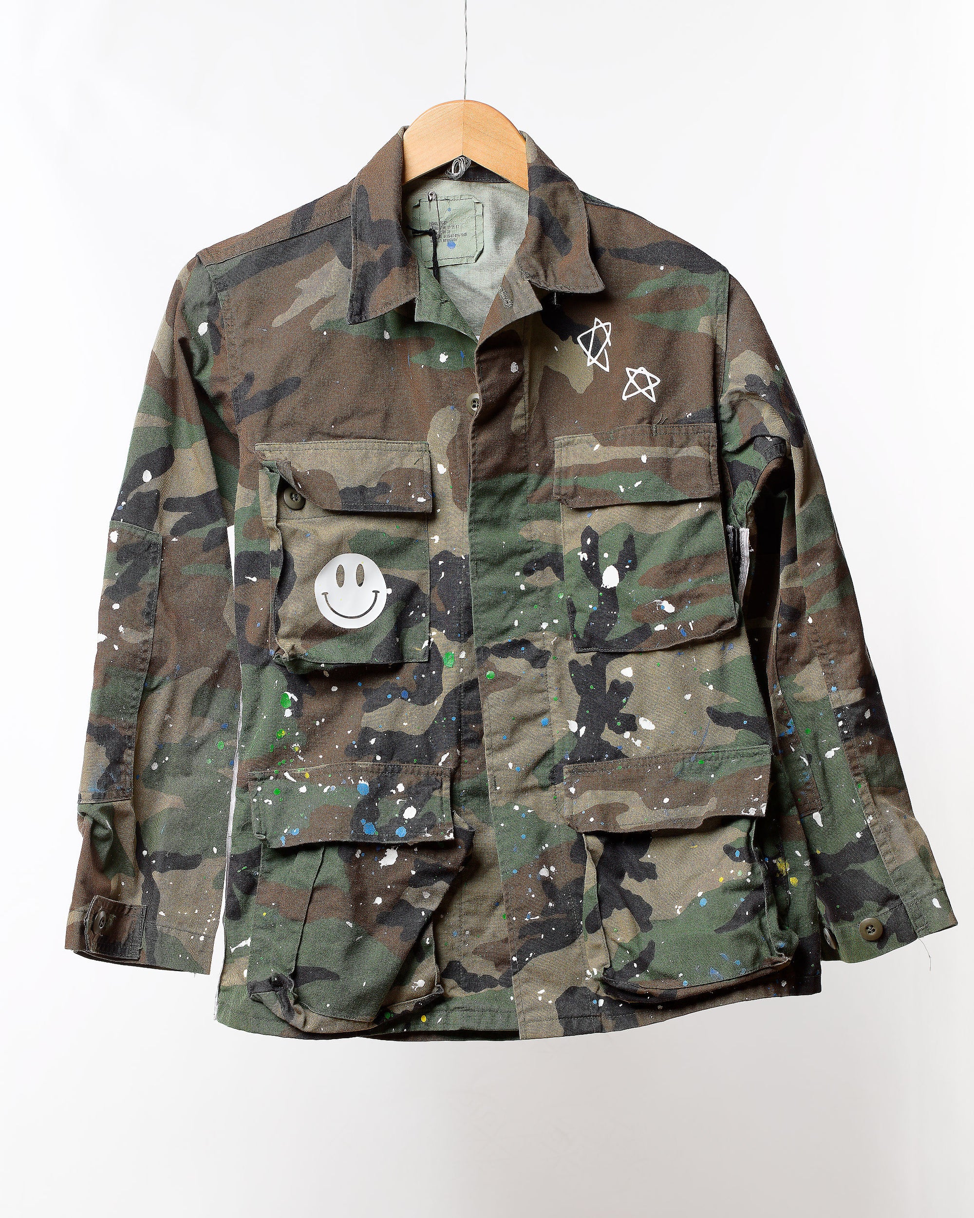 The Tak Design "Camo Jacket with Splatter & Trim" features a vintage military style adorned with colorful paint splatters, a smiley face patch on one pocket, and star drawings on the chest. This unique fashion piece is displayed on a wooden hanger against a white background.