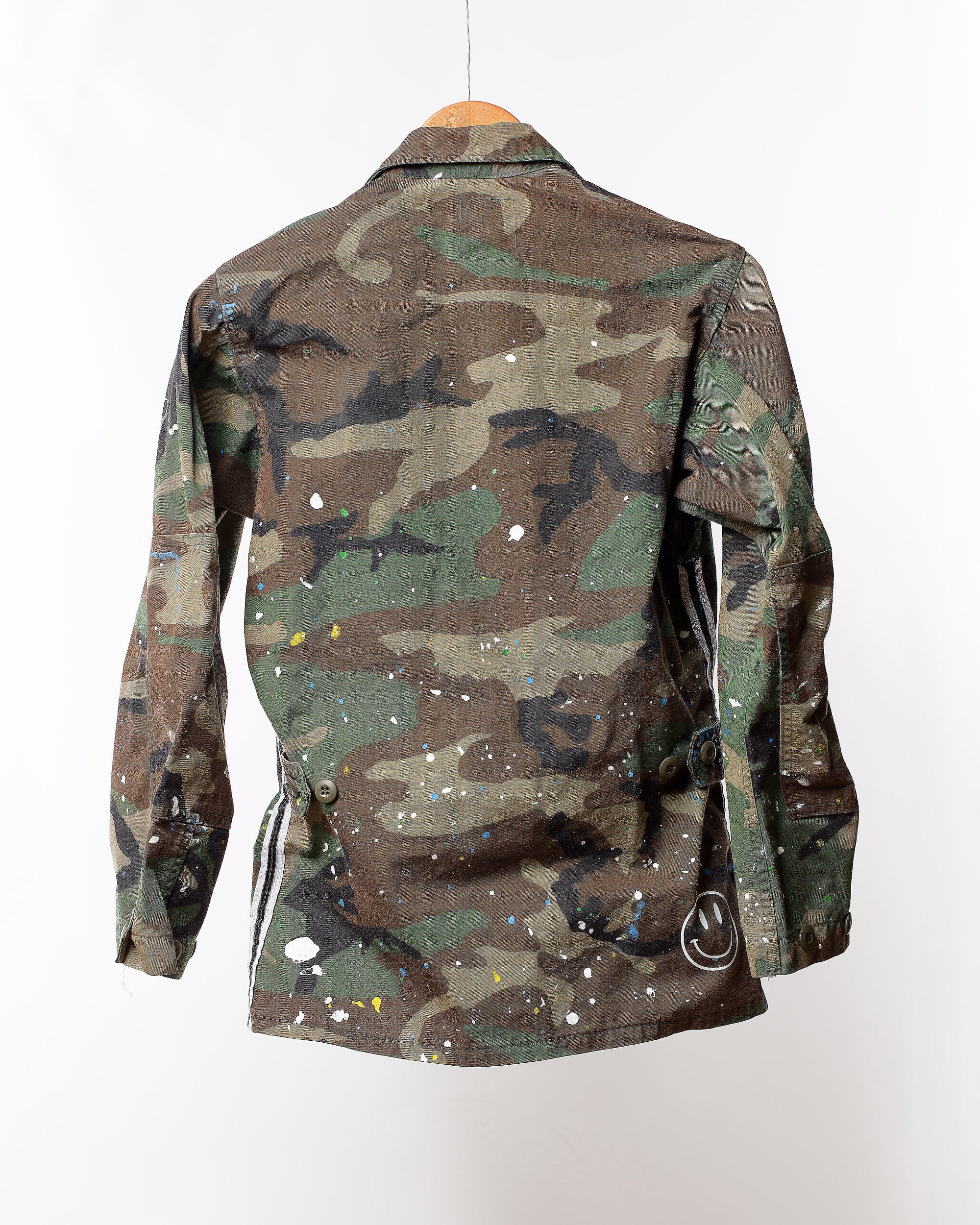 A Camo Jacket from Tak Design hangs on a wooden hanger against a plain white background. This unique fashion piece, named the "Camo Jacket with splatter & Trim," is artfully adorned with white, yellow, and black paint splatters, offering an artistic and modern aesthetic.