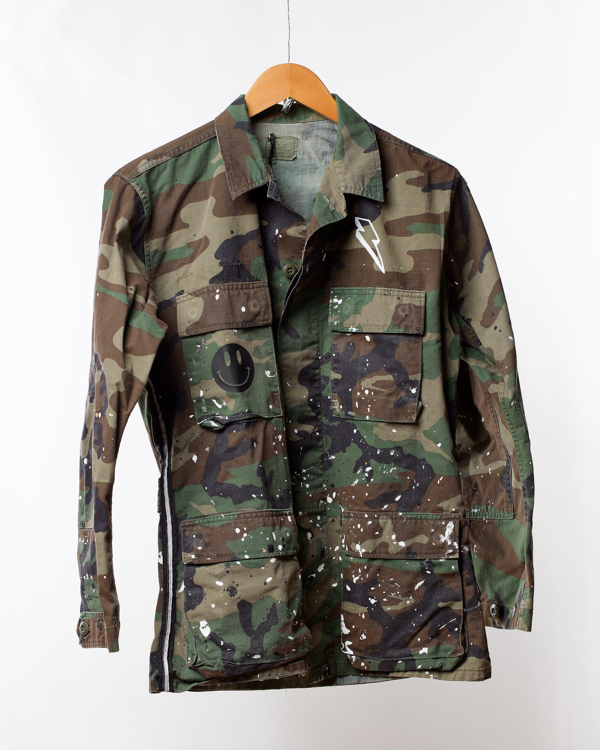 The "Camo Jacket with splatter & Trim" from Tak Design is displayed on a hanger, featuring a black smiley face badge and a lightning bolt patch. Its unique fashion details include white paint splatters that accentuate the camouflage pattern in shades of green, brown, and black.