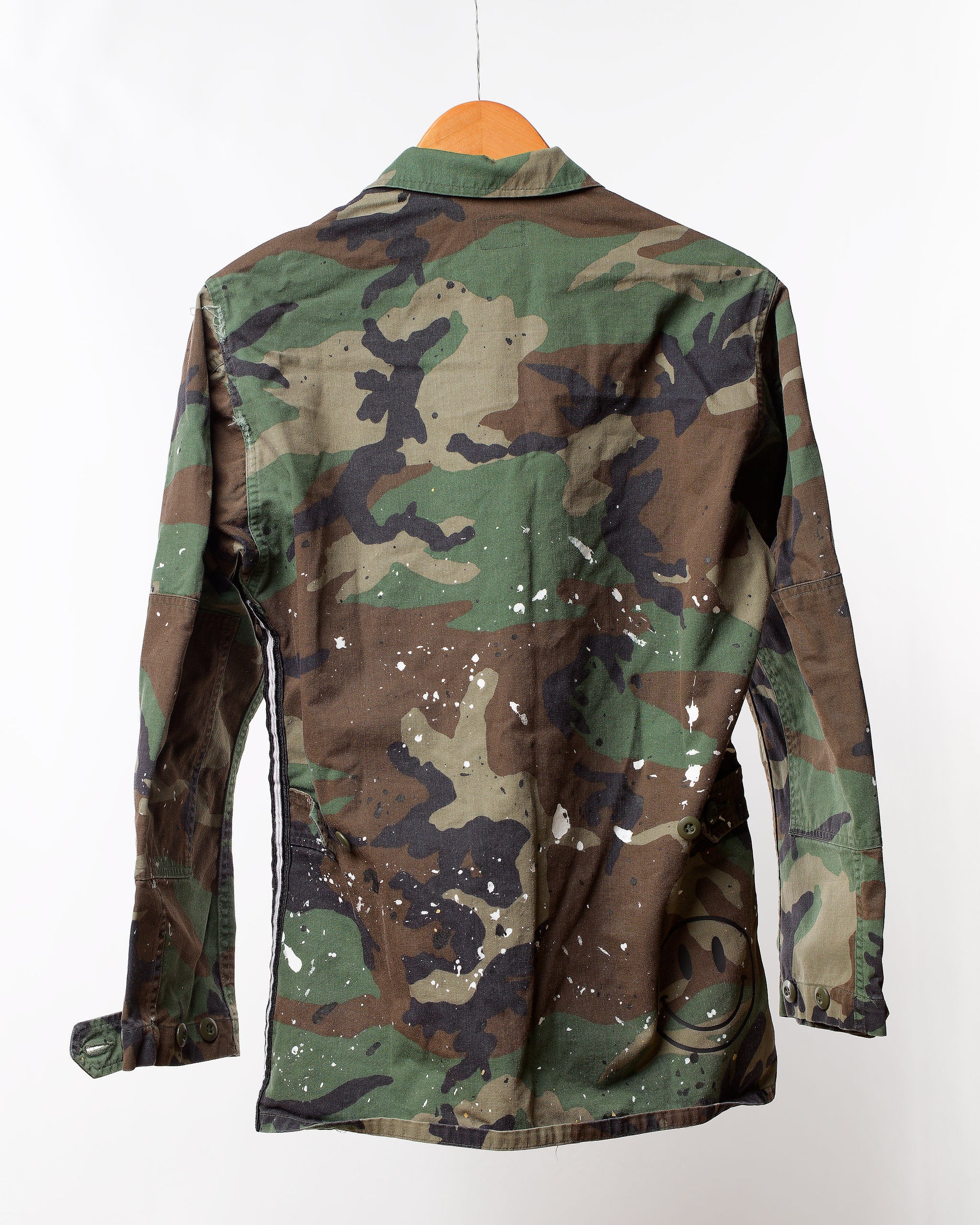 A Camo Jacket with splatter & Trim by Tak Design hangs on a wooden hanger, showcasing its camouflage fabric accented with white paint splatters. A black smiley face grins from the bottom right corner, adding a rugged, worn charm to this distinctive fashion piece.