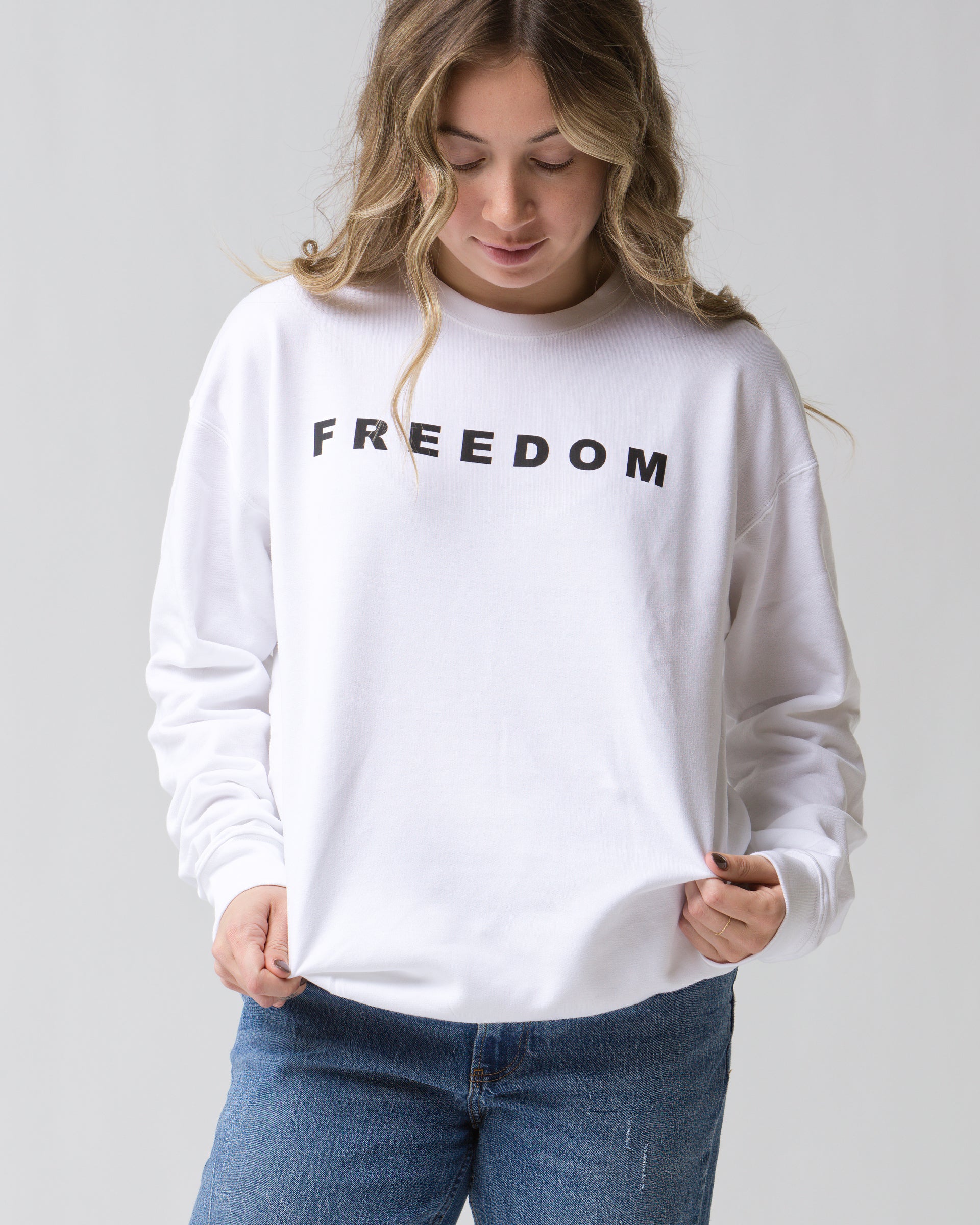 A person in the Faire FREEDOM Charlie Kirk Sweatshirt, featuring bold black "FREEDOM" lettering, stands against a plain background looking down and softly holding the hem—capturing conservative activist Charlie Kirk's spirit.