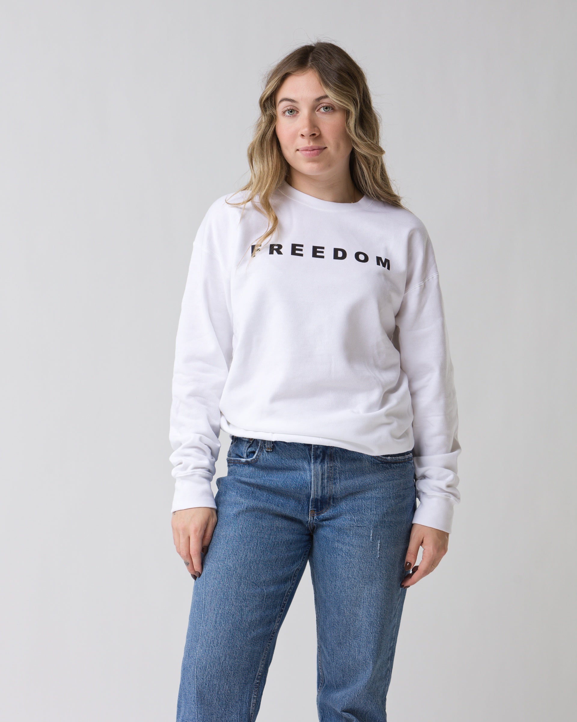A woman with wavy blonde hair stands against a plain background, wearing the Faire FREEDOM Charlie Kirk Sweatshirt and blue jeans. She looks ahead with a calm expression, capturing the vibe of a Turning Point USA conservative activist.
