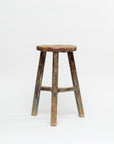 Against a plain white background stands the ROUND STOOL by Indus Design Imports, a worn, rustic wooden stool featuring four legs and a round seat. This imported handmade vintage piece showcases signs of age with patches of faded paint and scratches, exuding timeless elegance and weathered charm.