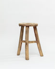 A handmade vintage ROUND STOOL 48 with timeless elegance, featuring a rustic wooden seat and four sturdy legs from Indus Design Imports. The stool boasts a worn, natural wood finish, giving it a well-used appearance that highlights its artisan craftsmanship. The background is plain white.