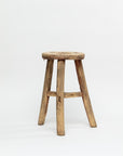 The ROUND STOOL by Indus Design Imports is a weathered, rustic wooden stool featuring four legs and a round seat. Showcasing timeless elegance, the stool's surface reveals natural wood texture and imperfections, giving it a worn, antique appearance. This piece of artisan craftsmanship stands against a plain white background.