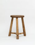 The ROUND STOOL by Indus Design Imports showcases a rustic wooden design featuring a round seat and four sturdy legs. This stool exudes timeless elegance with its natural, unfinished appearance that beautifully highlights the wood's texture and grain. The artisan craftsmanship is evident in the horizontal braces connecting the legs, providing added stability. Photographed against a plain white background, this piece stands out for its simplicity and charm.