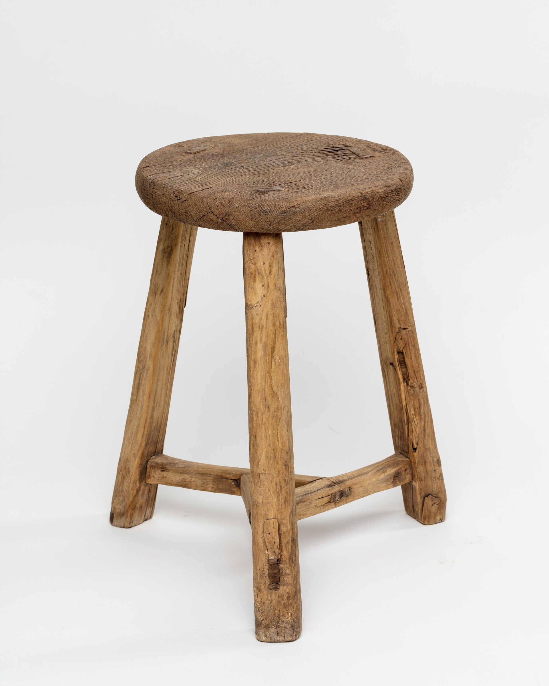 The ROUND STOOL by Indus Design Imports features a rustic wooden design with a round, worn seat and three slightly angled, sturdy legs connected by a triangular support structure. The stool&#39;s visible wood grain and imperfections showcase artisan craftsmanship, giving it a timeless, handmade appearance against a plain white background.