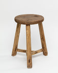 The ROUND STOOL by Indus Design Imports features a rustic wooden design with a round, worn seat and three slightly angled, sturdy legs connected by a triangular support structure. The stool's visible wood grain and imperfections showcase artisan craftsmanship, giving it a timeless, handmade appearance against a plain white background.