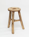 Introducing the ROUND STOOL by Indus Design Imports, a rustic piece crafted from wood with three legs and an unfinished, rough surface. Showcasing artisan craftsmanship, this stool features visible grain patterns and natural imperfections in the wood, giving it a weathered and vintage appearance. It stands alone against a plain white background.