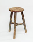 The ROUND STOOL by Indus Design Imports is a rustic, weathered wooden stool featuring a round seat and three legs. Showcasing timeless elegance, the stool is aged with worn paint and visible signs of use, reflecting its vintage charm. This piece of artisan craftsmanship stands out beautifully against a plain white background.