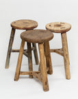 Three ROUND STOOLs by Indus Design Imports, featuring round, weathered seats and sturdy legs, are arranged on a plain white background. Displaying signs of wear and aging, these handmade vintage stools showcase variations in wood color and texture, emphasizing their timeless elegance and artisan craftsmanship.