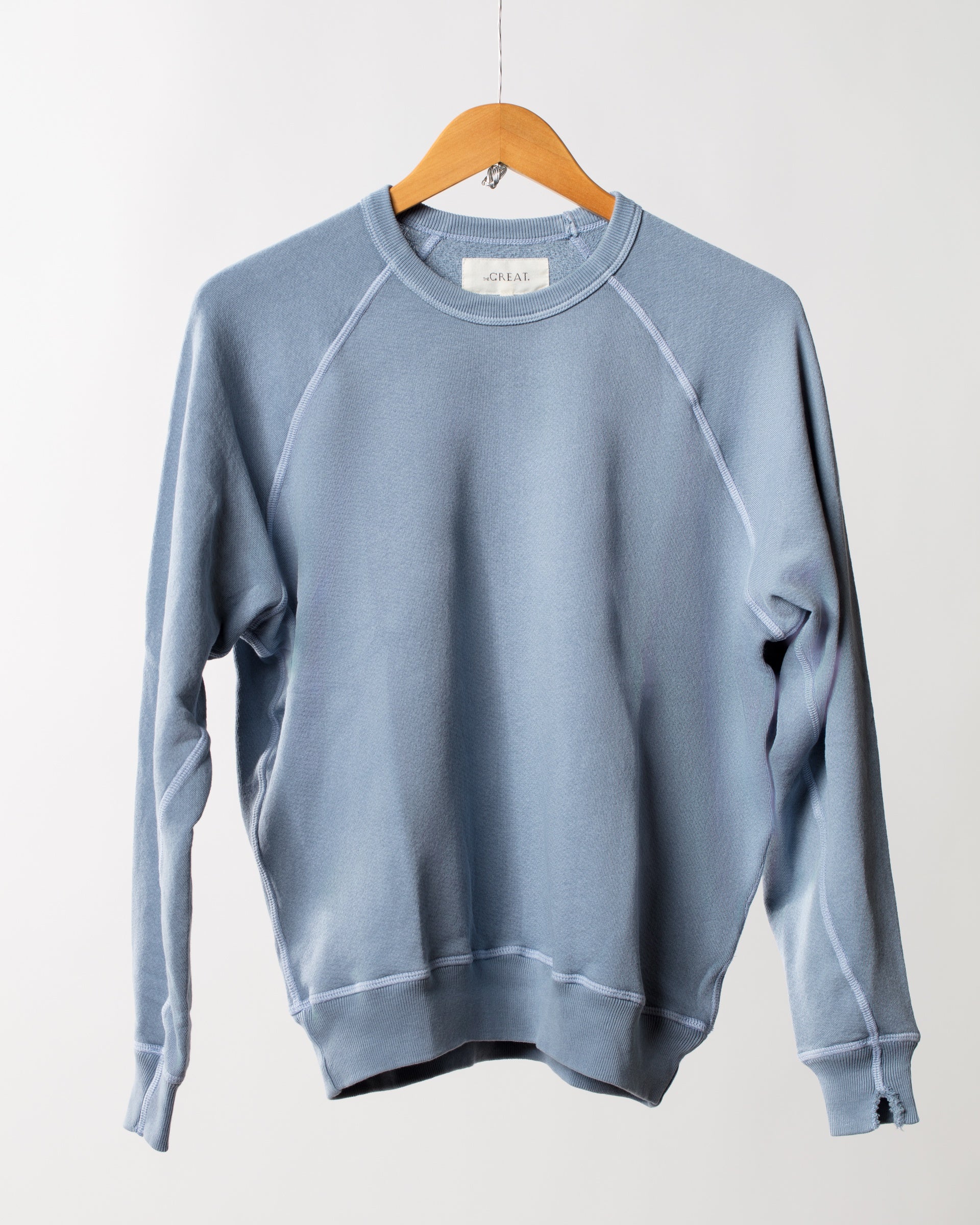 A light blue crewneck, The College Sweatshirt BGSKY by The Great Inc., hangs on a wooden hanger against a plain white background. This sweatshirt features long sleeves with visible seams and a ribbed hem and cuffs, creating a casual and comfortable look.