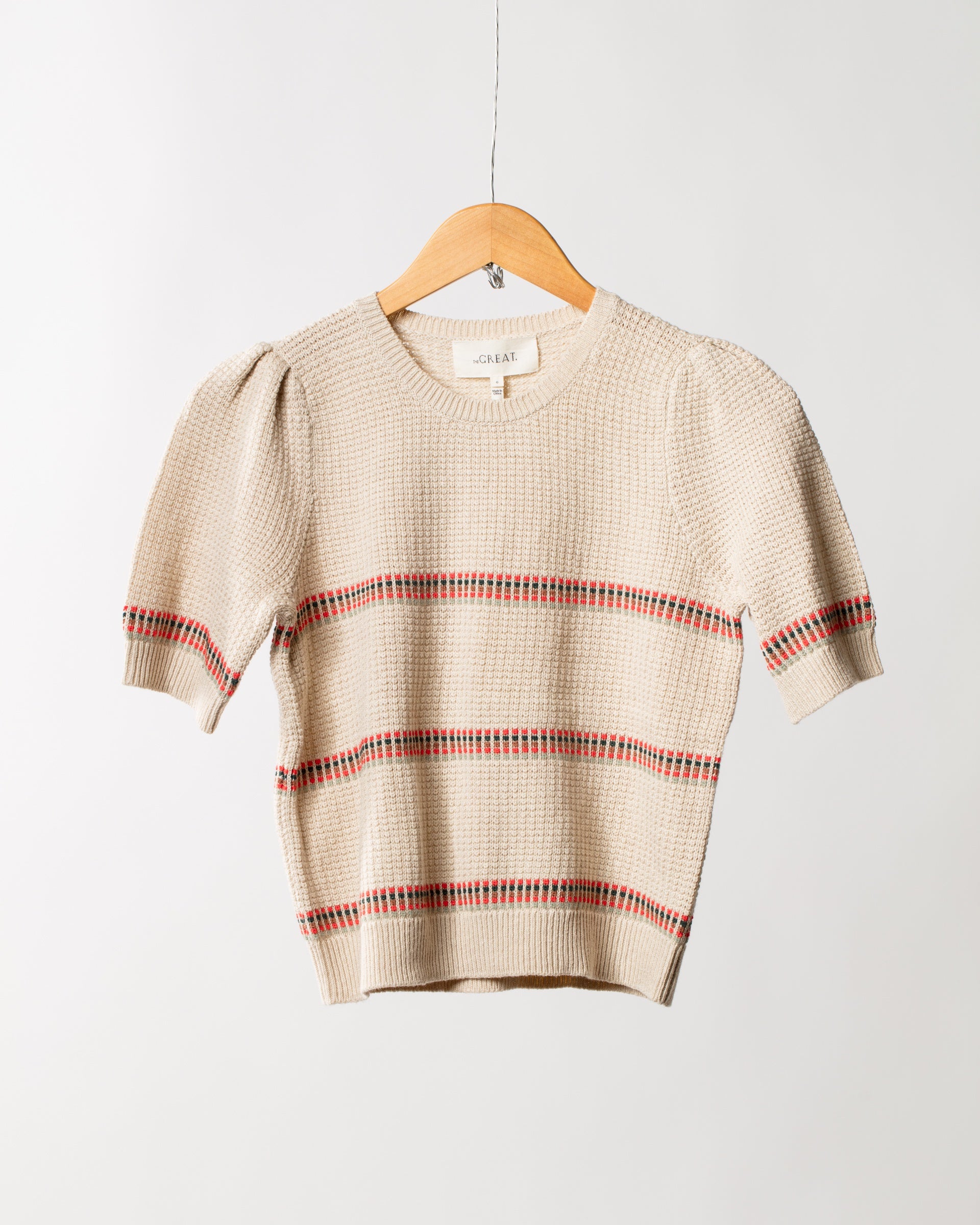 A beige knitted short-sleeve pullover from The Great Inc., named The Puff Sleeve Pullover, featuring red and black horizontal stripes, hangs on a wooden hanger against a white background. The pullover showcases a ribbed texture and puffed shoulders.