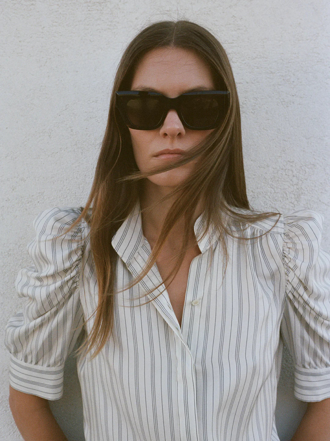 A woman with long hair and sunglasses wears the Gillian Top by FRAME, featuring a white, striped design with cotton ruched puff sleeves as she stands against a textured, light-colored wall. Her expression is neutral, complementing the timeless elegance of her attire.