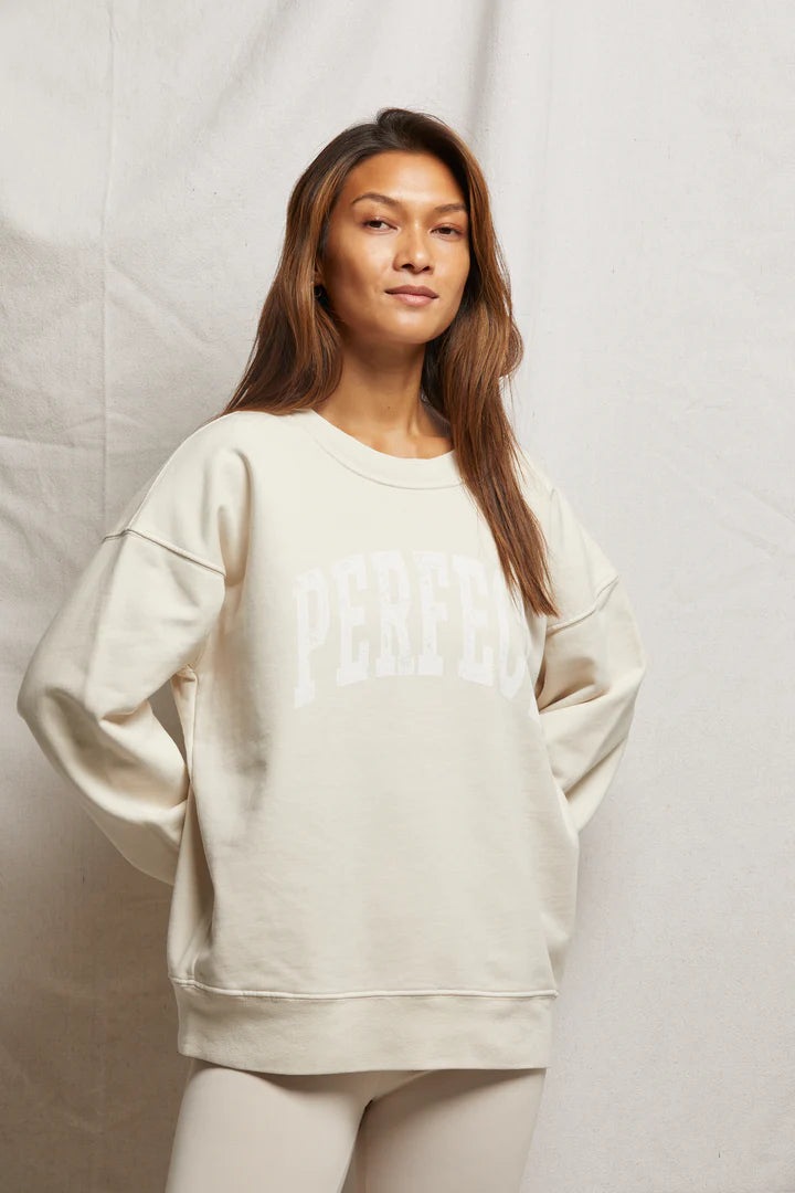 A person stands wearing a cream-colored Perfect Sweatshirt by Perfectwhitetee, featuring "Perfect" in varsity letters. With long light brown hair and a slight smile, they face forward. The oversized fit pairs well with the plain background's light hues.