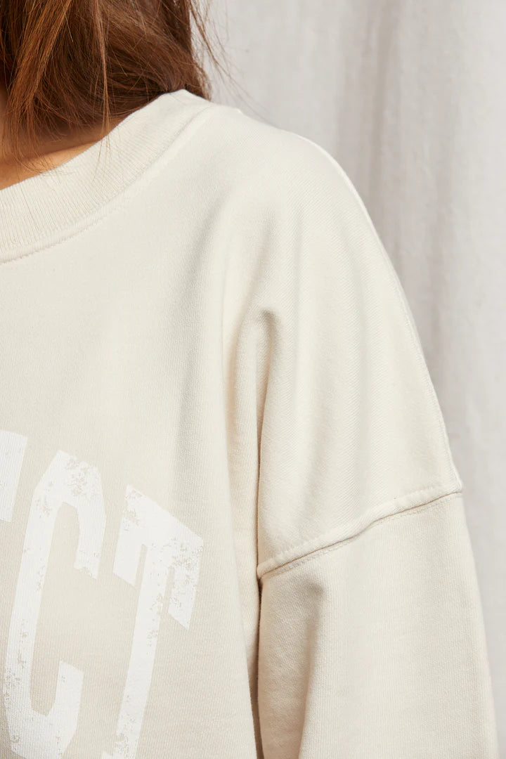 A close-up of the Perfectwhitetee's cozy, oversized Perfect Sweatshirt showcases its cream hue and partially visible white varsity letters against a plain, neutral backdrop.