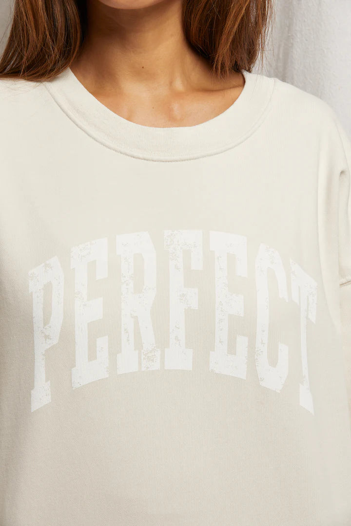 Close-up of a person wearing the Perfectwhitetee's Perfect Sweatshirt, an oversized beige crewneck with "PERFECT" in large, slightly distressed varsity letters across the chest, focusing on the upper section.