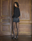 A woman with long curly hair stands before a wooden paneled wall, wearing the DOEN Petra Crystal Cardigan, black satin lace-trim shorts, patterned tights, and black heels. She glances over her shoulder at the camera.