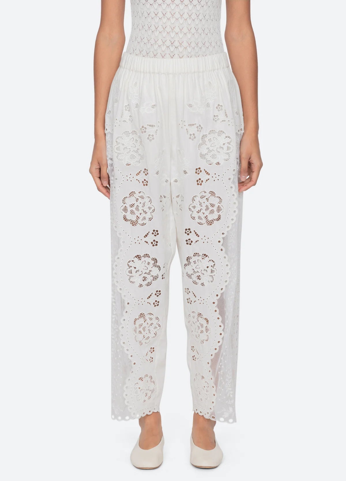A person wearing Sea New York Gardener Pants—vintage-inspired sheer white pants with floral cut-out embroidery and an elastic waistband—paired with white flats stands against a plain light background.