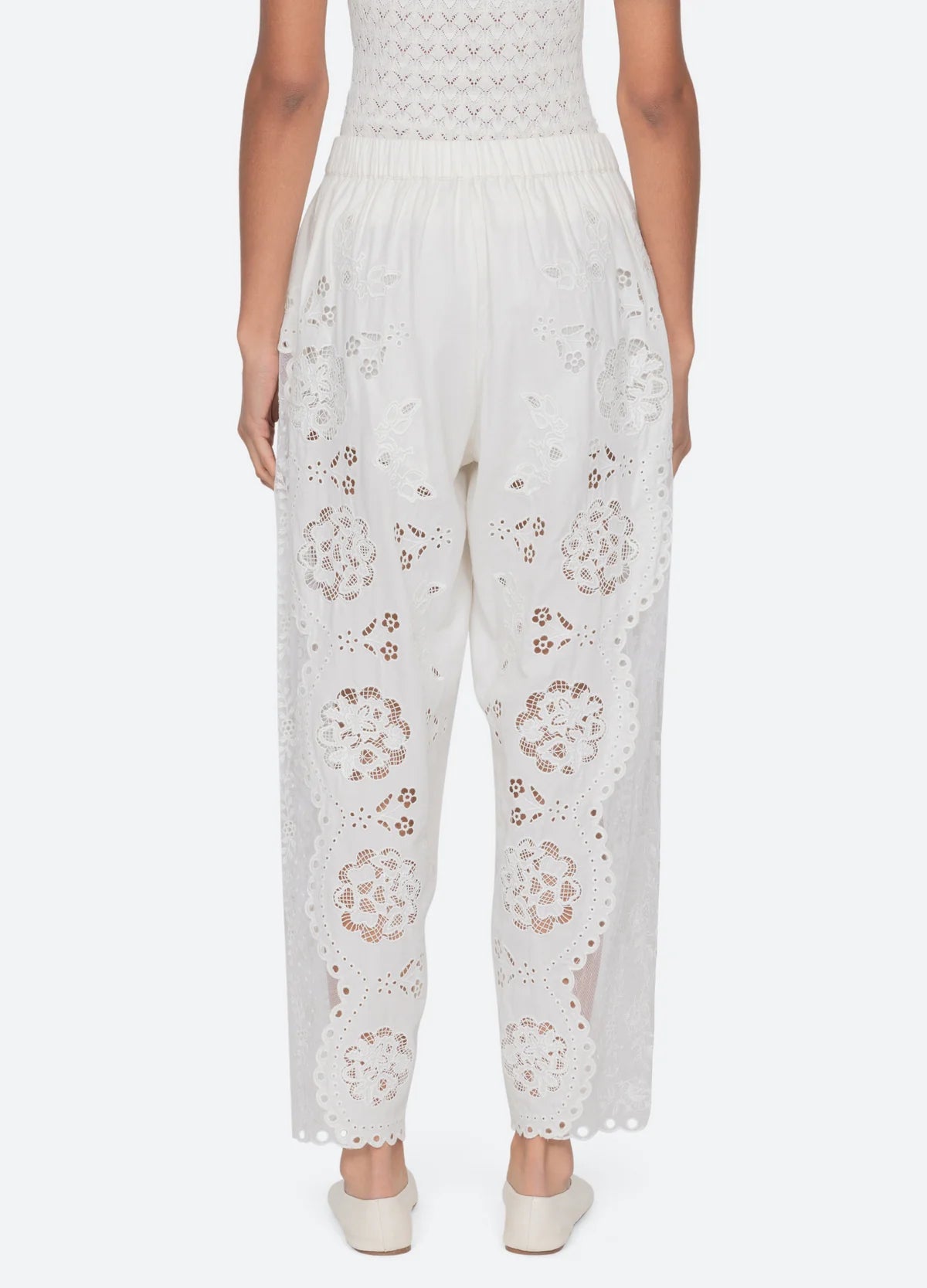 A person faces away wearing Sea New York Gardener Pants—vintage-inspired, loose white pants with eyelet lace and scalloped edges—paired with white slip-on shoes and a textured white top.