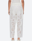 A person faces away wearing Sea New York Gardener Pants—vintage-inspired, loose white pants with eyelet lace and scalloped edges—paired with white slip-on shoes and a textured white top.
