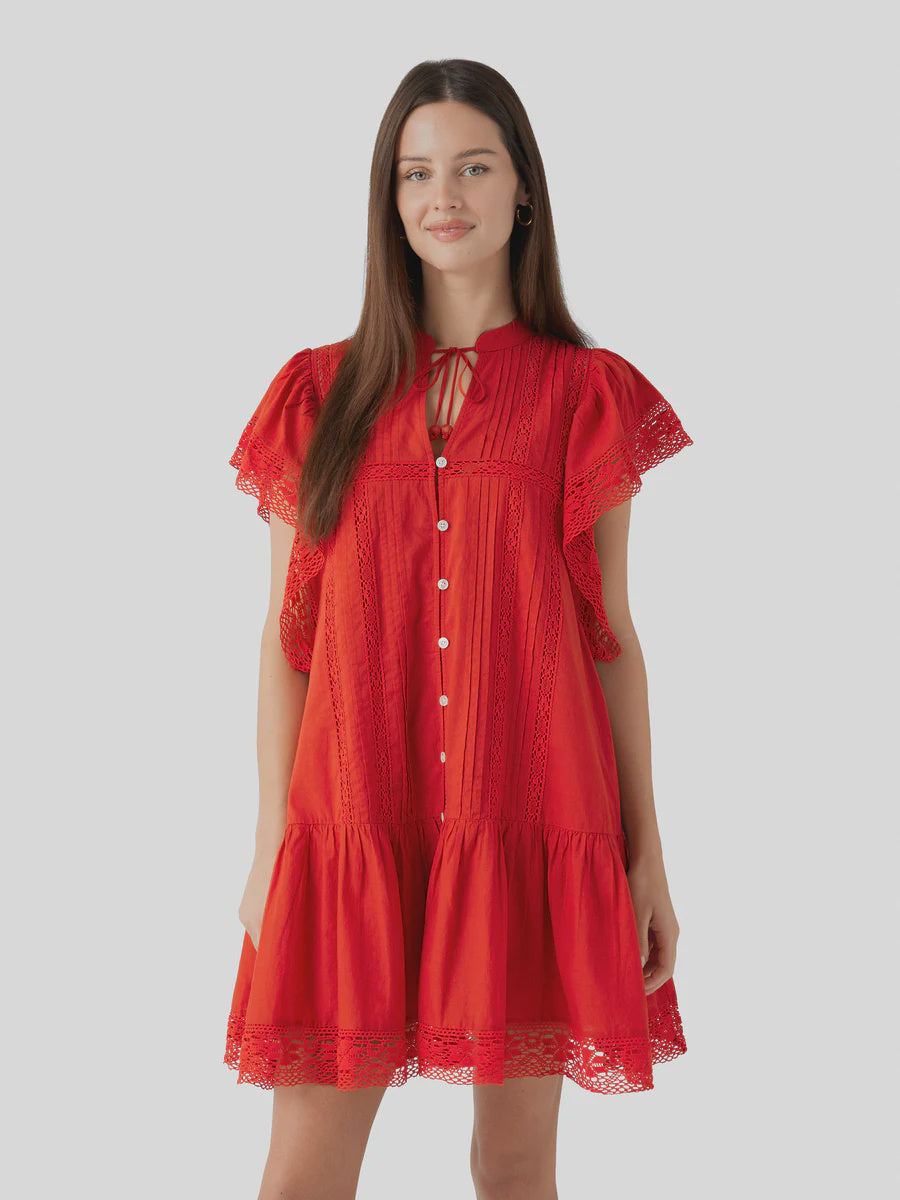 A woman smiles gently while wearing the Sea New York Pippa SS Mini Dress, a vintage-inspired bright red mini dress with lace details, flutter sleeves, and front button-down design, standing against a plain light gray background.