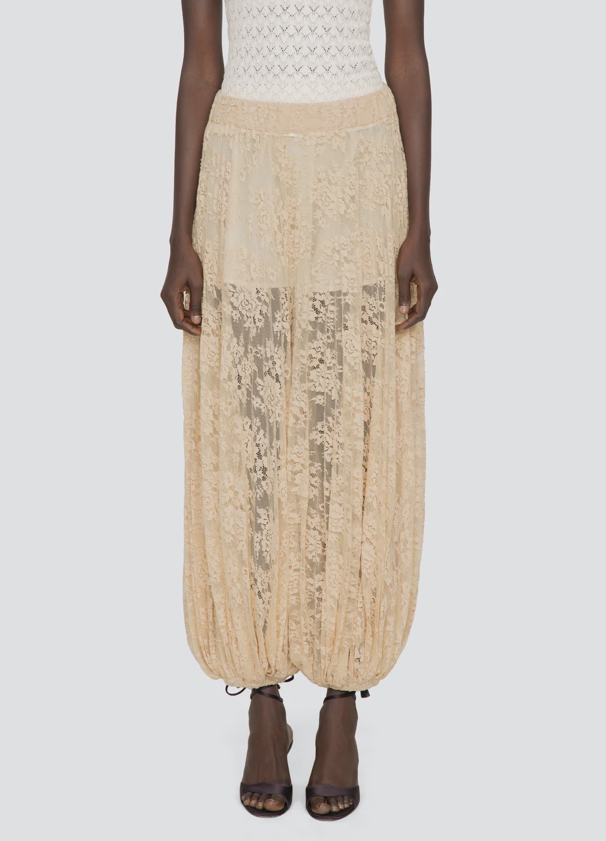 A person wears the Sharleen Lace Pant by Sea New York—a beige floral lace, sheer balloon-silhouette pant with gathered hems—styled with a white textured top and black open-toe heels against a plain light background.