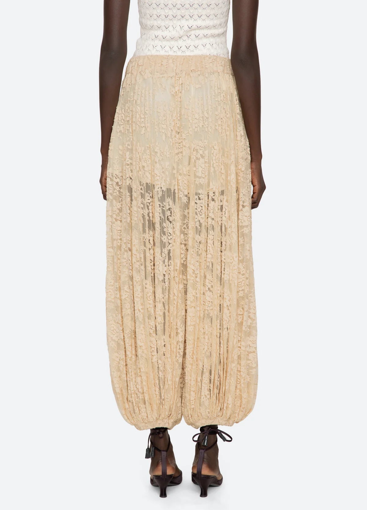 A person shown from the back wears the Sea New York Sharleen Lace Pant—sheer beige floral lace with a balloon silhouette and gathered ankles—paired with black heeled sandals, against a plain light background.