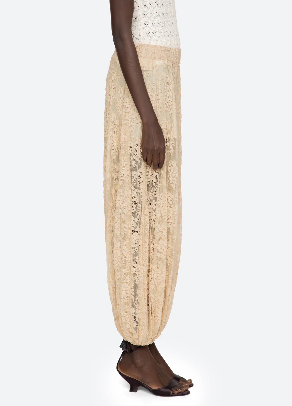 A person wearing the Sea New York Sharleen Lace Pant in light beige, styled with a white sleeveless top and black heeled sandals, stands in profile against a plain background.