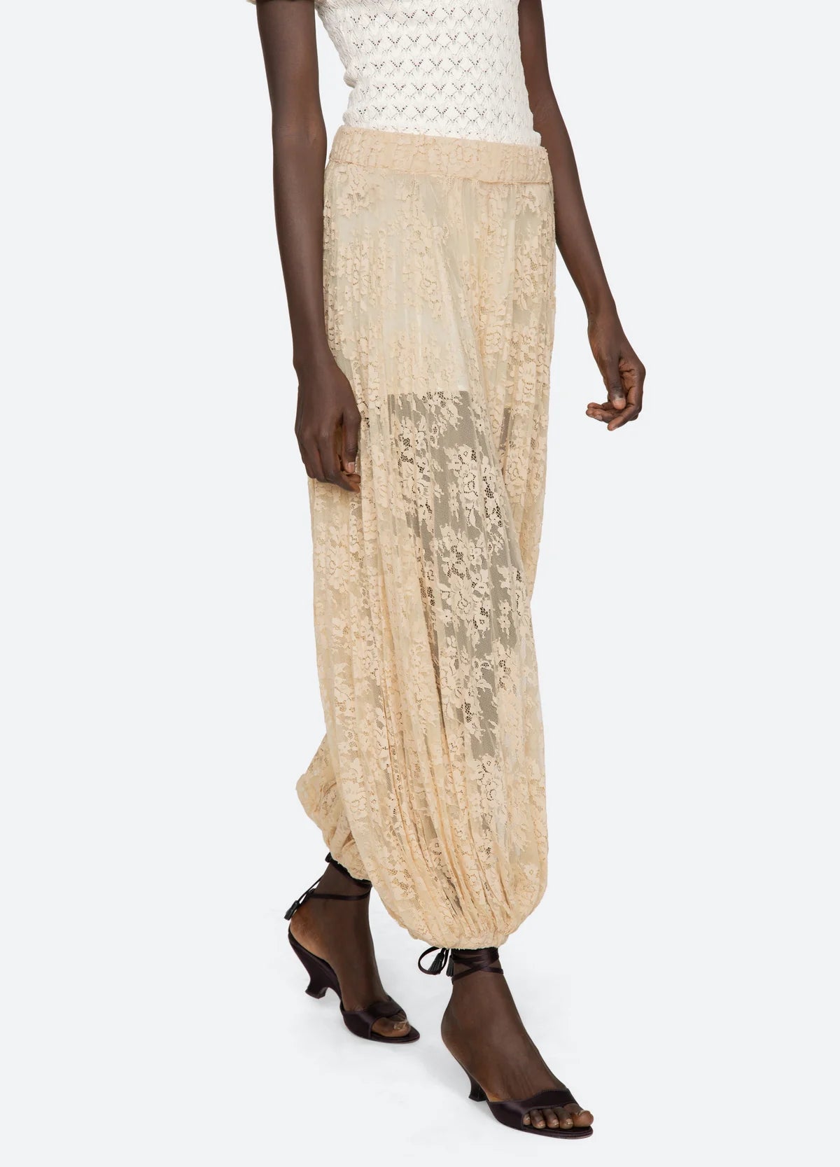 A person wears the Sharleen Lace Pant by Sea New York—sheer beige floral lace with a balloon silhouette, elastic waistband, and gathered ankles—paired with a white textured top and black heeled sandals.